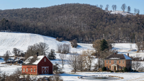Photograph of a rural landscape with a stately brick farmhouse surrounded by manicured landscaping, a small pond in the foreground, and a traditional red barn with a cupola to the left. Rolling green hills and a dense forest form the background under a clear blue sky.