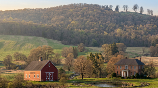 Photograph of a rural landscape with a stately brick farmhouse surrounded by manicured landscaping, a small pond in the foreground, and a traditional red barn with a cupola to the left. Rolling green hills and a dense forest form the background under a clear blue sky.
